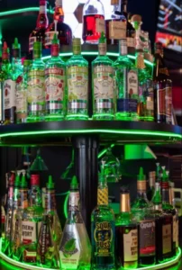 The image shows the cozy interior of Chicago Pub Cambridge with green lighting illuminating the shelves filled with bottles. The bar area is lively with patrons enjoying their drinks, contributing to the warm and welcoming atmosphere.