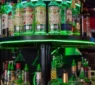 The image shows the cozy interior of Chicago Pub Cambridge with green lighting illuminating the shelves filled with bottles. The bar area is lively with patrons enjoying their drinks, contributing to the warm and welcoming atmosphere.