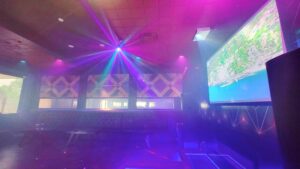 The image shows an interior of a lounge or club with colorful lights, a large screen displaying a map, cushioned benches, and tables, creating a vibrant and inviting atmosphere.